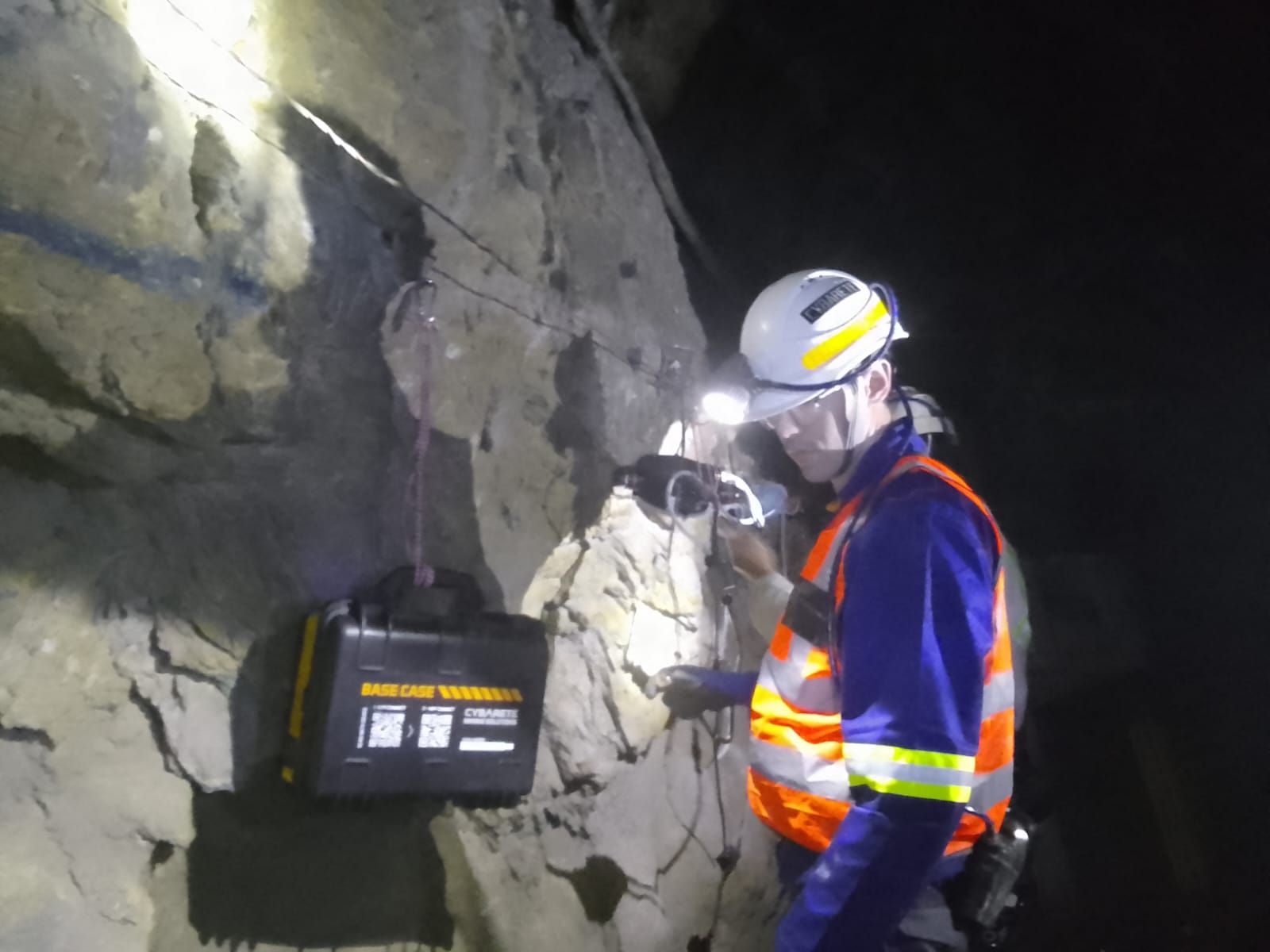A miner in a hard hat and reflective vest works at the rock face beside a BASE Case hung on the sidewall.