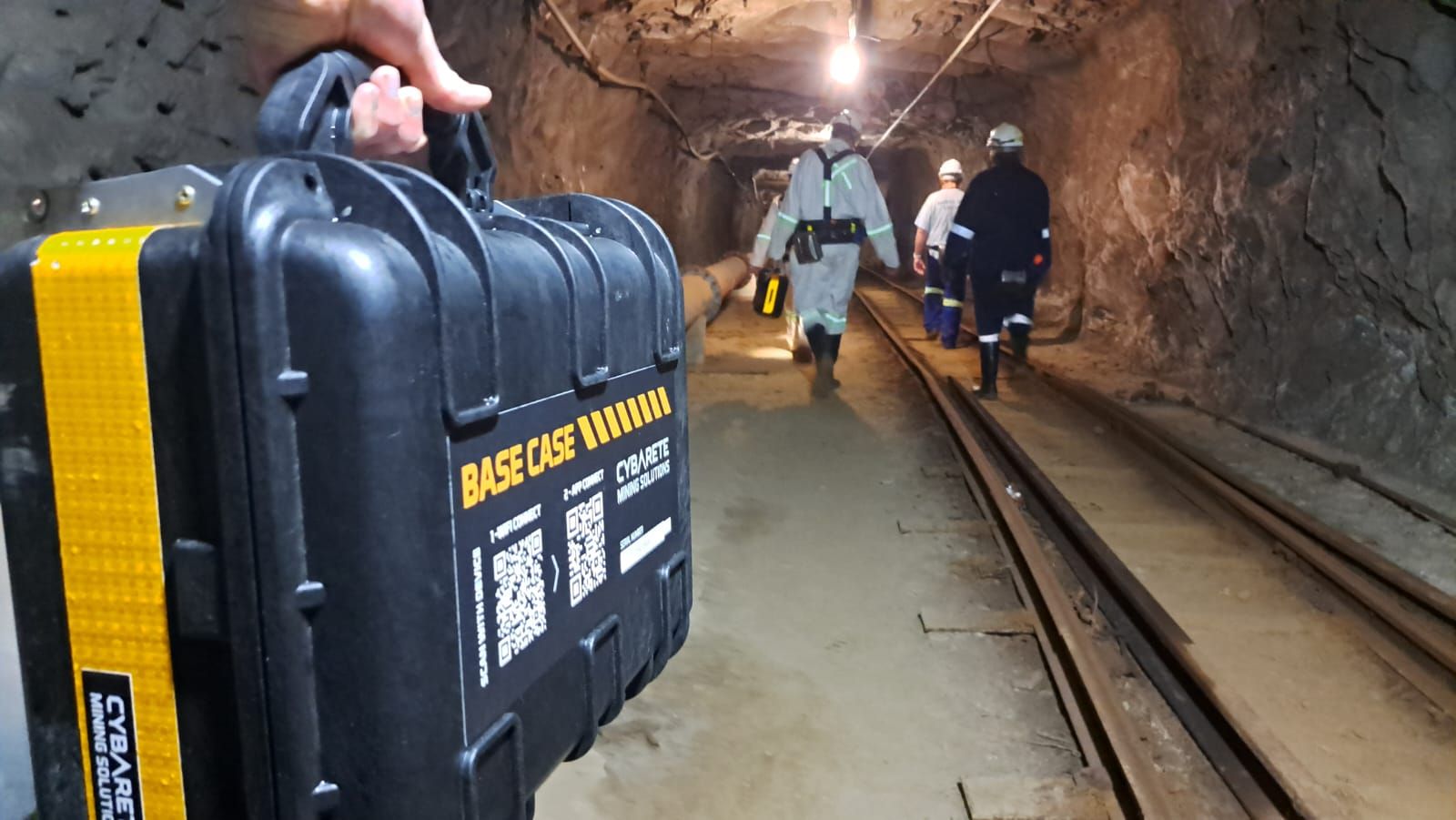 A BASE Case carried by its handle along an underground haulage, with miners walking ahead beside the rail track.